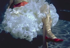 A woman is seen from waist down, sitting in a white ruffled dress, with orange belt. Her legs are crossed, with one briliant red shoe showing. She may be underwater or just behind glass with water highlights, reflected onto her.