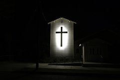 A cross is shillouetted with strong light. It is centered on a rectangular wall, with peaked roof. A wheelchair ramp is in front, leading to a doorway.