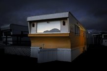 A trailer home is centered in the composition. It has a window reflecting mountains with lamps on each side of the window. The trailer has an orange band around the center, white on top and a white fence around the bottom. The sky is dark.