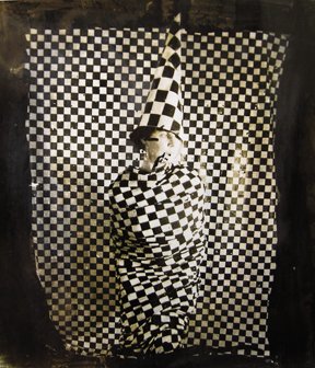 A checkered sheet is behind a figure wrapped in a checked sheet with a dunce hat, also in black and white squares.