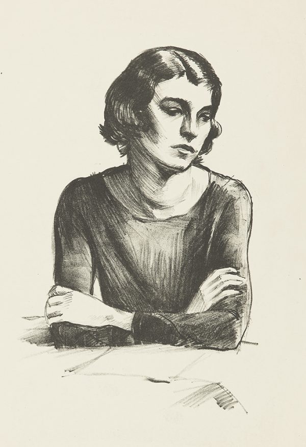 A woman leans on a table with arms crossed. She has an open paper in front of her but looks off to her left.
