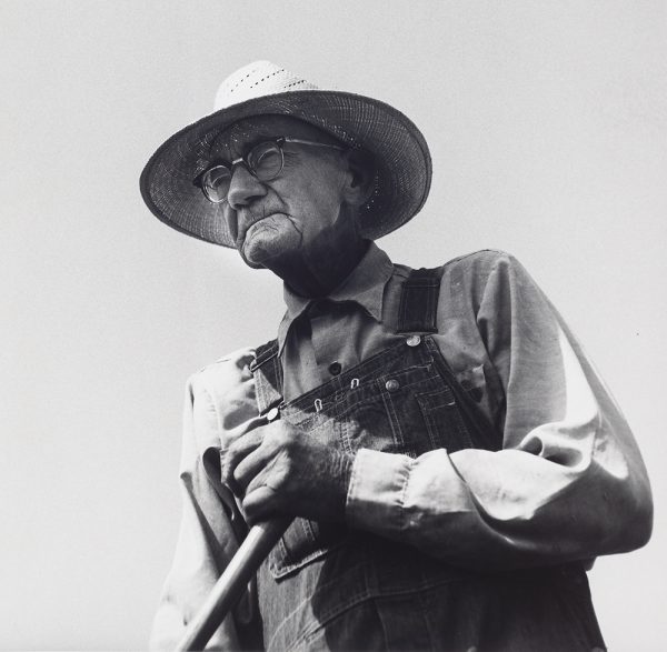 Old man in hat and glasses in overalls with yard utensil.