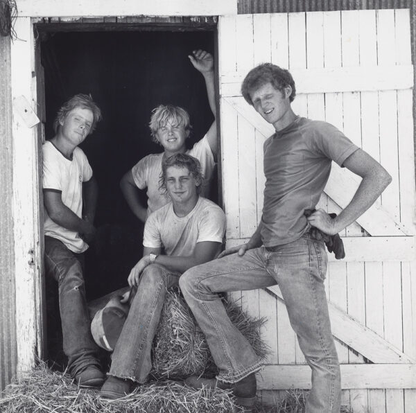 Four sweaty young men in frame of white painted barn door. One sitting on bail of hay, holding a hat, one behind him resting proper left arm up on the frame of the door, proper right hand on his hip, one leaning on the left side of the door frame, one standing by the door with proper right foot resting on the bail of hay, holding gloves at his waist. All wearing T-shirts, jeans, and boots.