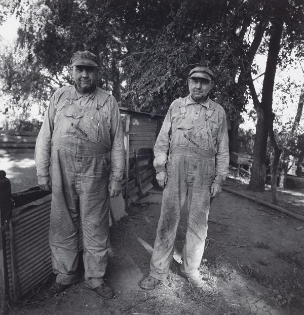Two men stand on dirt ground near fence wearing hats and overalls.