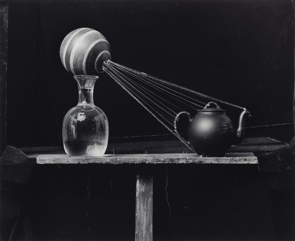 Sitting on a small wooden table, a glass vase holds a stripped ball with strings attached at one end. These strings are attached at the other end to a small, dark teapot to the right.