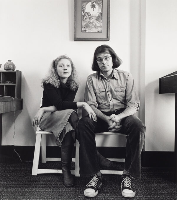 Young couple sits together posing in wooden chairs against a plain wall with a framed image above them. They look at the camera and do not smile.