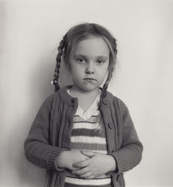 Young girl with pig tail braids holds her hands in front of her and poses against plain wall. She looks at camera with no smile.