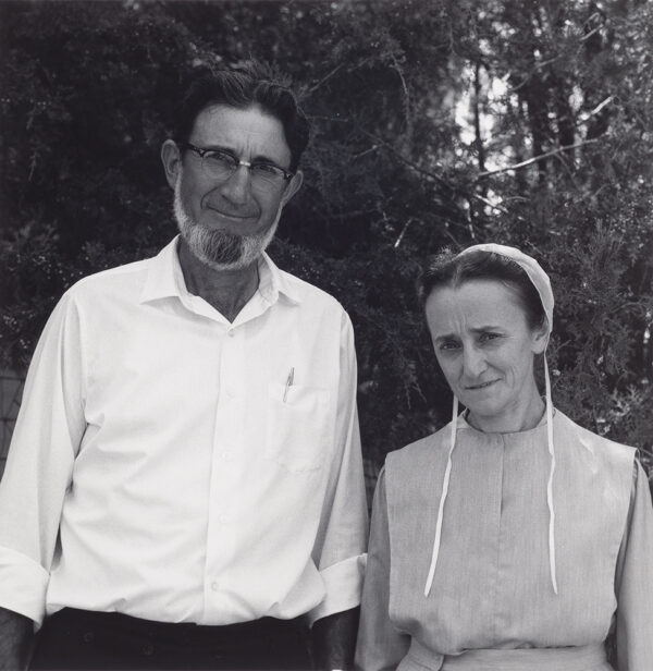Conservative couple stands in front of trees. The man has a beard and wears a white shirt. The woman wears a white bonnet.