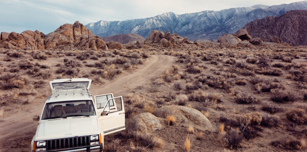 White Jeep car with both proper left doors and trunk open is pulled over on a rocky desert landscape background with mountains in the distance.
