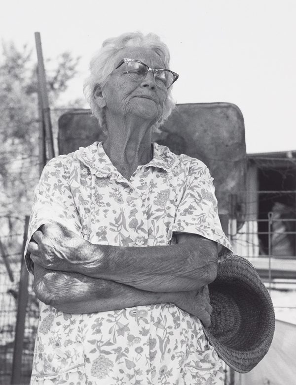 Elderly woman with glasses and arms crossed holding a hat in proper right hand.