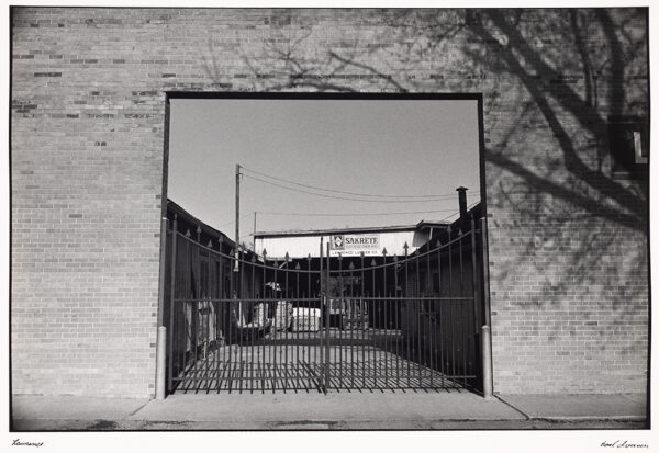 A brick wall with a fenced off square opening to a Lawrence lot with a sign reading 