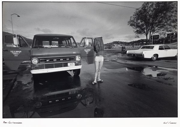 Woman stands in the street of a parking lot, pulling skirt over her head to expose undergarments and argil knee socks. Next to her is a Ford van with 