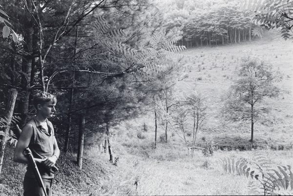 Young man stands in a field with forest behind him. He holds a shotgun in his proper right arm.