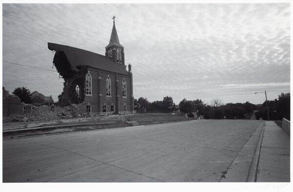 A half torn down church stands at the left with a street in front of it, There are trees in the background and clouds encompassing the entire sky.