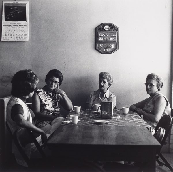 Four older women sit at two wooden tables, pushed together, one covered with table cloth. Each woman has a coffee cup in front of them and a napkin dispenser in the center of the table. Plaque on wall  above table reads 