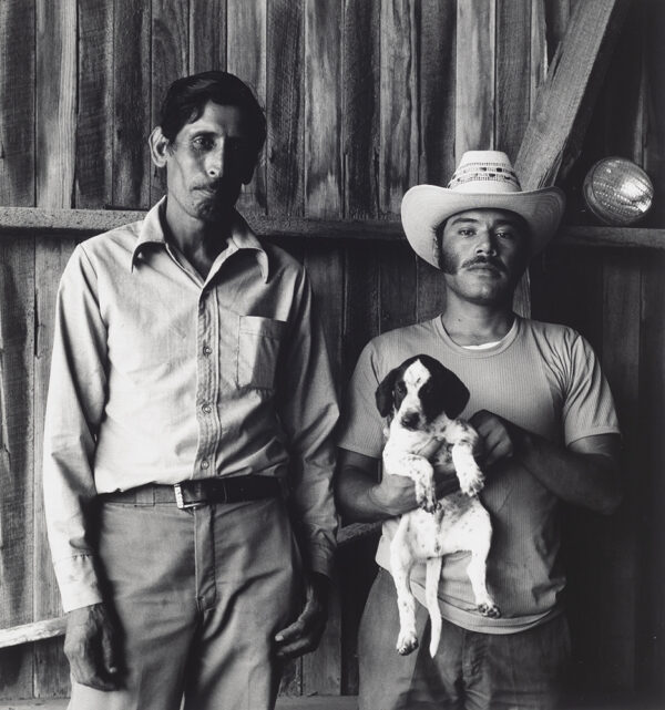Two men standing with a wooden barn background. The man on the right, wearing a hat, holds a small, black and white dog.
