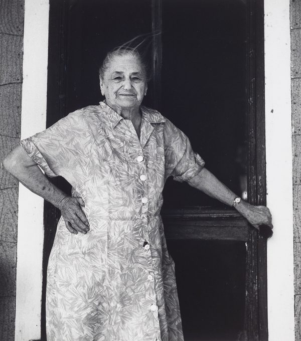 Older woman standing at screen door with proper right hand resting on hip.