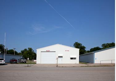 Front view of building with a large roll up door that is set back from the street. Two adjacent white building are partially visible. Three cars are parked next to the left building. Trees are popping above the roofs. Blue sky is clear with a white line of smoke, as evidence of a plane passing by.