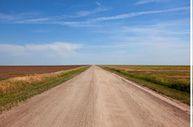 One-point perspective with flat horizon line. Blue sky, dirt road, grass growing along the sides, barren fields.