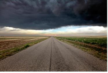 A very dark storm cloud hovers over a dry landscape with a road running down the center.