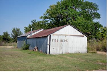 Green grass, white fire department building with rust brown roof, trees in the background