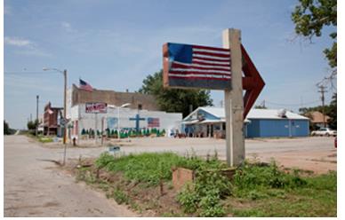 American flag painted on a wooden post in the foreground, patch of green grass. Buildings and streetlights with flags flying in the background.