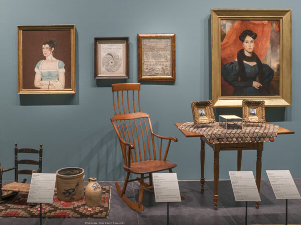 Photo of early American decorative arts includes two chairs, one rocking chair, two paintings of somen, a desk, framed photos, a rug, a milk jub, a crock and a duck decoy