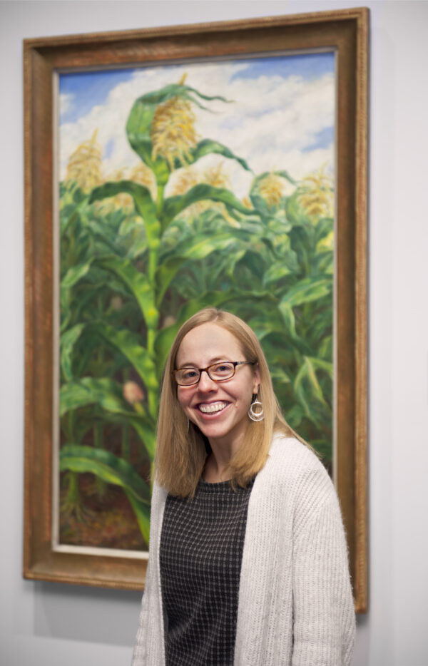Woman wearing a grey sweater and blue dress in front of a painting of a cornfield with green stalks and yellow tassles
