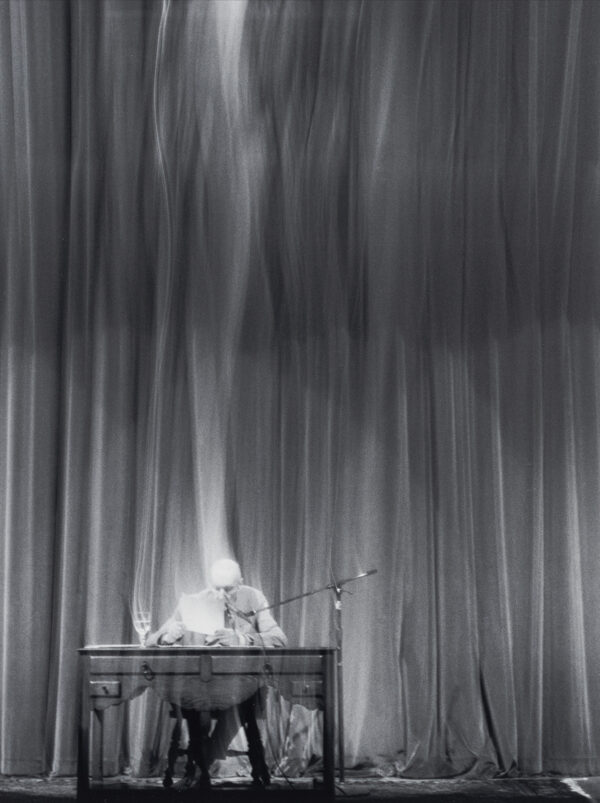 William S. Burroughs giving a reading at the Wichita Center for the Arts; he is seated at a table on stage in the theatre with a curtain behind him.