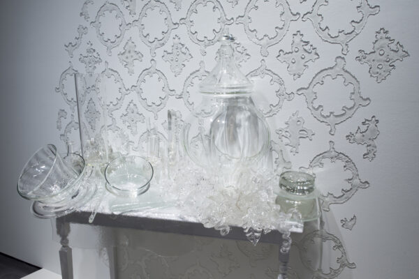 Detail of a clear glass sculpture of vases and other vessels laid on a white table with ornate glass wallpaper on the wall behind the table
