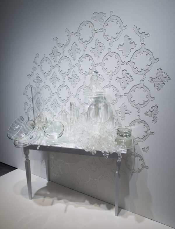 White wooden table covered in glass objects, forms and containers of varying sizes with glass decor attached the wall behind