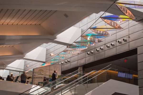 Interior photo of Wichita's Eisenhower Airport with a large-scale multicolored artwork hanging in sections from the ceiling over an escalator with visitors
