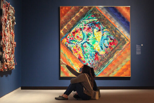 Photo of two people sitting on the floor in front of a colorful abstract painting, with one pointing to another painting on the all in front of them.