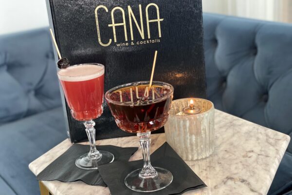 Cana Wine and Cocktails Menu behind two mixed drinks on a whiite lace tablecloth