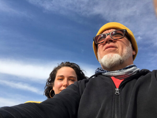 Photograph of a bearded man with glasses and weaing a knitted yellow cap with a woman with dark hair and weaing large gold hoop earrings looking over his shoulder