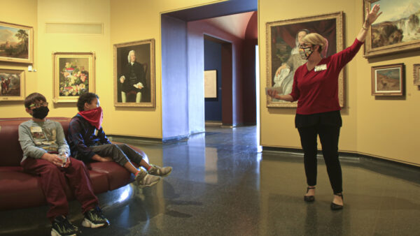 Interior of the galleries with a woman on the right gesturing toward a painting and two young men on the left sitting and watching