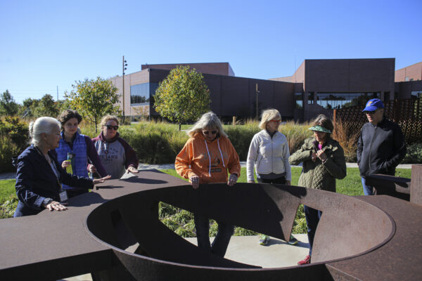 Photograph of a group of people looking at a large-scale circular sculpture of weathered metal in the museum art garden with the museum building in the background