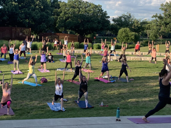 Photo of four dozen people in colorful yoga clothing with mats and water bottles on the grass lawn space in the Downing Amphitheater participating in a yoga class with arms outstretched above their heads