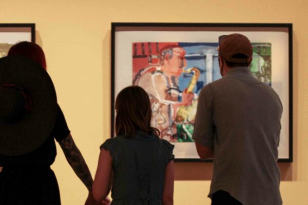 Three people, adult woman holding the hand of a young girl and adult man, in silhouette in front of colorful painting of a saxophone player in a gallery