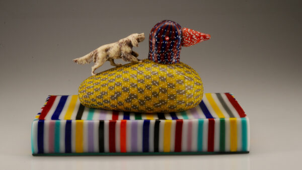 Colorful glass base with duck on top of it and a dog on the left climbing up the back of the duck