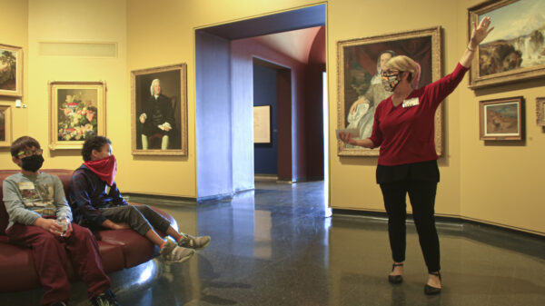 Woman on the right in the galleries gesturing towards a painting while two boys on the left sit on a round sofa and watch