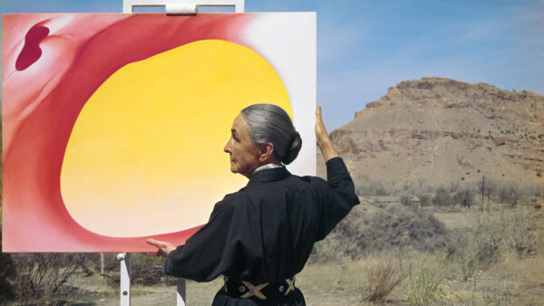 Photograph of Georgia O'Keeffe holding a painting in front of a landscape of desert mountains