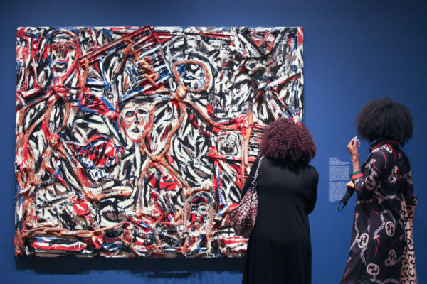 Two women with their backs toward the camera looking at a multi-colored abstract painting against a blue gallery wall