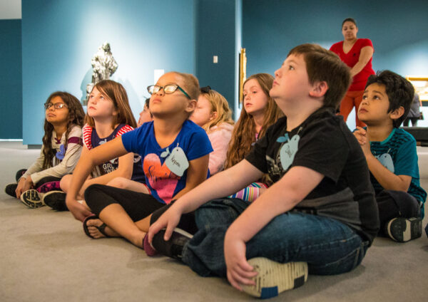 Group of students sitting on the floor in a gallery, looking up.