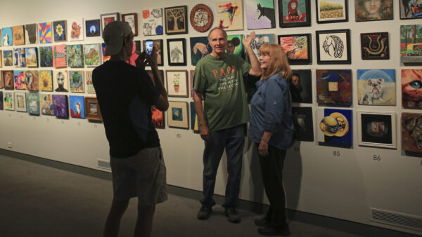 Two people standing in front of a wall of 12x12-inch paintings from the Foot in the Door exhibition while another visitor takes their photo