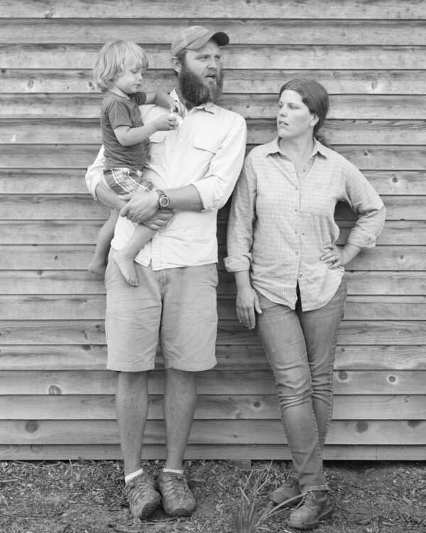 Black and white photograph of a man, holding a small child, and a woman, standing in front of a rough wood background
