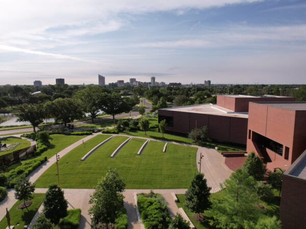Aerial photo of the Art Garden with green grass, limestone benches, the red brick museum building, trees in the front and the City of Wichita skyline in the background