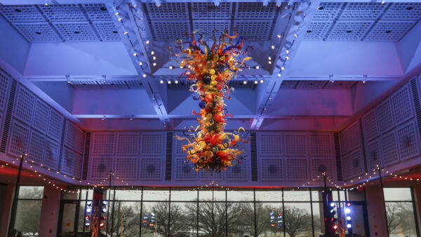 Photo of the colorful, large-scale Chihuly Chandelier in the S. Jim and Darla Farha Great Hall with small, white lights hanging from the ceiling around it and large windows in the background