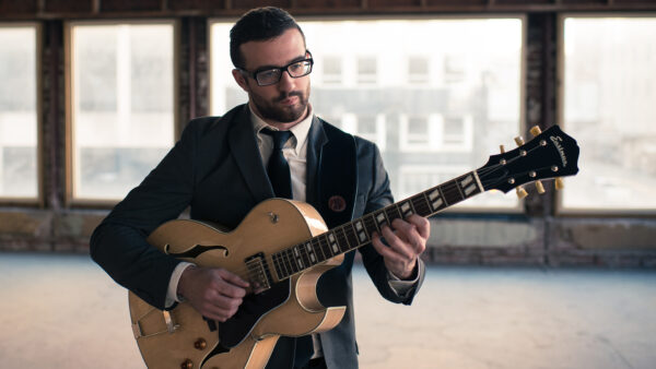 Photograph of a man dressed in a dark suit, white shirt and dark tie playing a guitar. He is standing in front of a bank of windows with a hazy urban landscape.