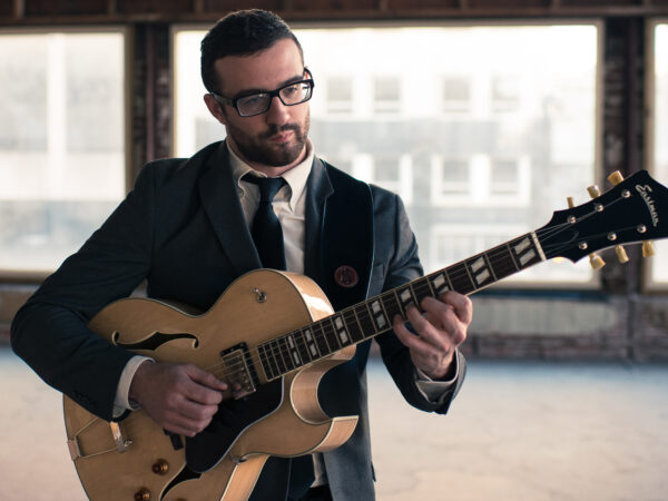 Photograph of a man dressed in a dark suit, white shirt and dark tie playing a guitar. He is standing in front of a bank of windows with a hazy urban landscape.